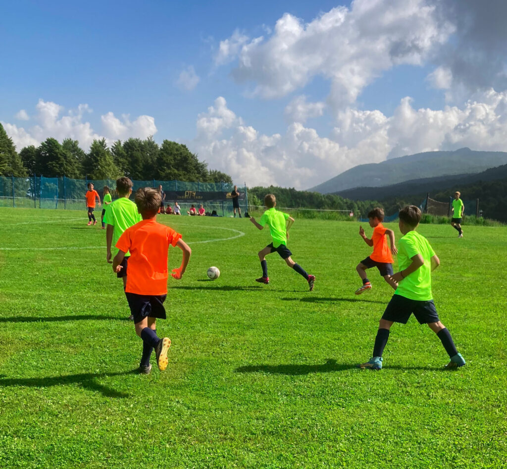Due squadre di ragazzi, con casacche verdi ed arancioni, che competono giocando a calcio su un campo presso Piancavallo.
